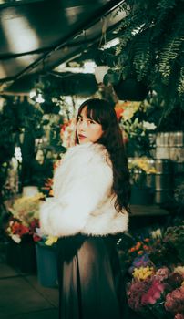 Elegant woman in a fur coat poses in a vibrant flower shop surrounded by rich greenery and colorful blooms.