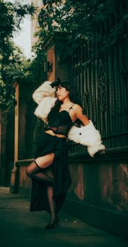 Chic woman posing in elegant black evening dress and white fur coat outdoors beside ornate fence.
