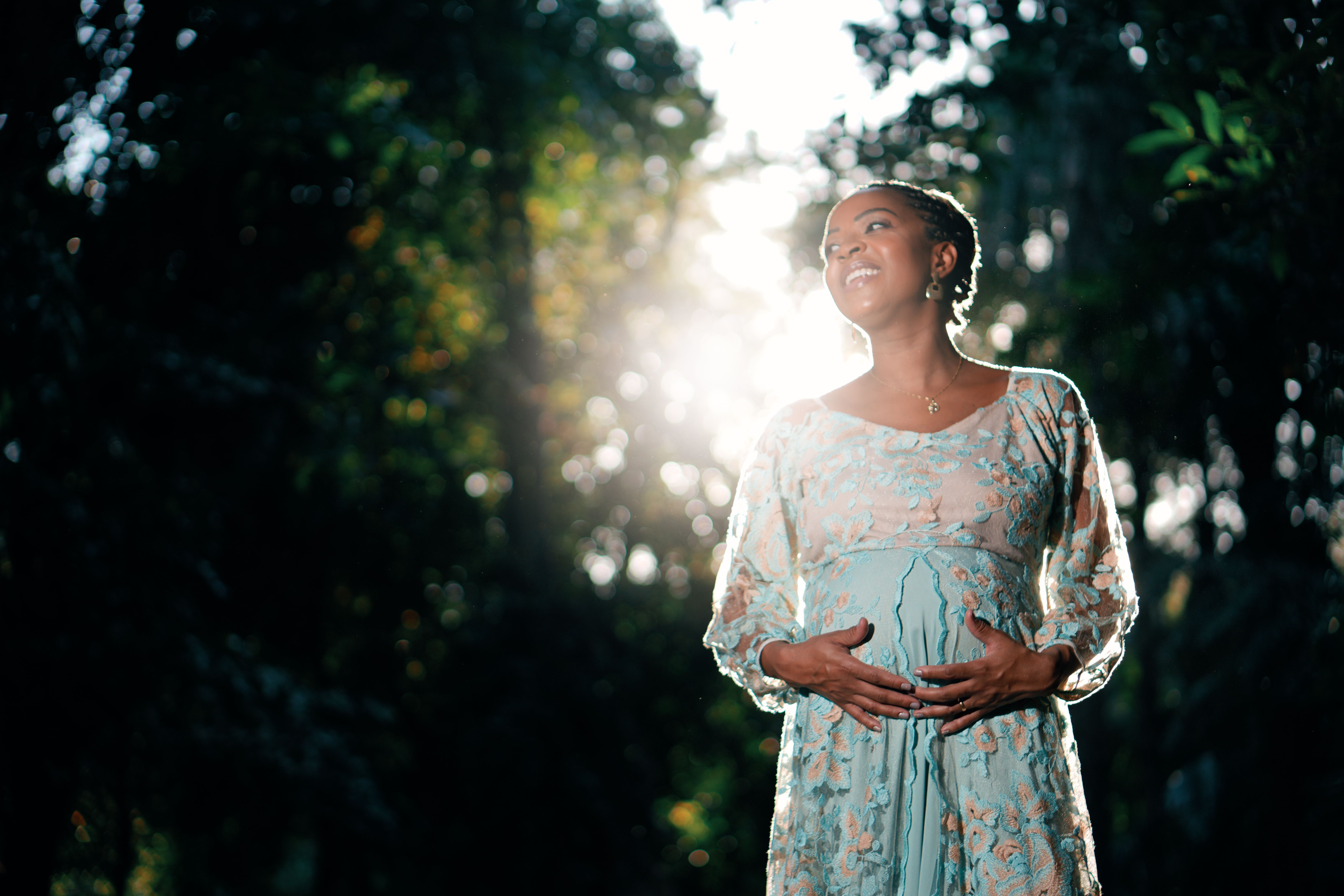 A serene pregnant woman in a sunlit garden wearing a floral dress.