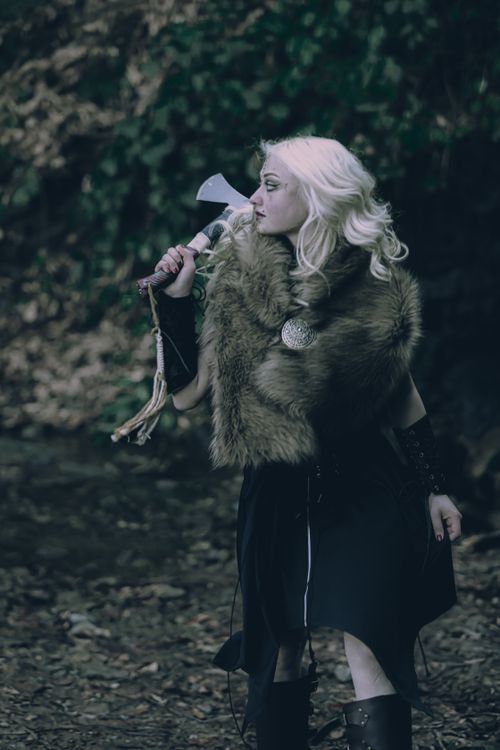 Free Blonde warrior woman in fur with axe in a dark forest setting, evoking a medieval fantasy era. Stock Photo
