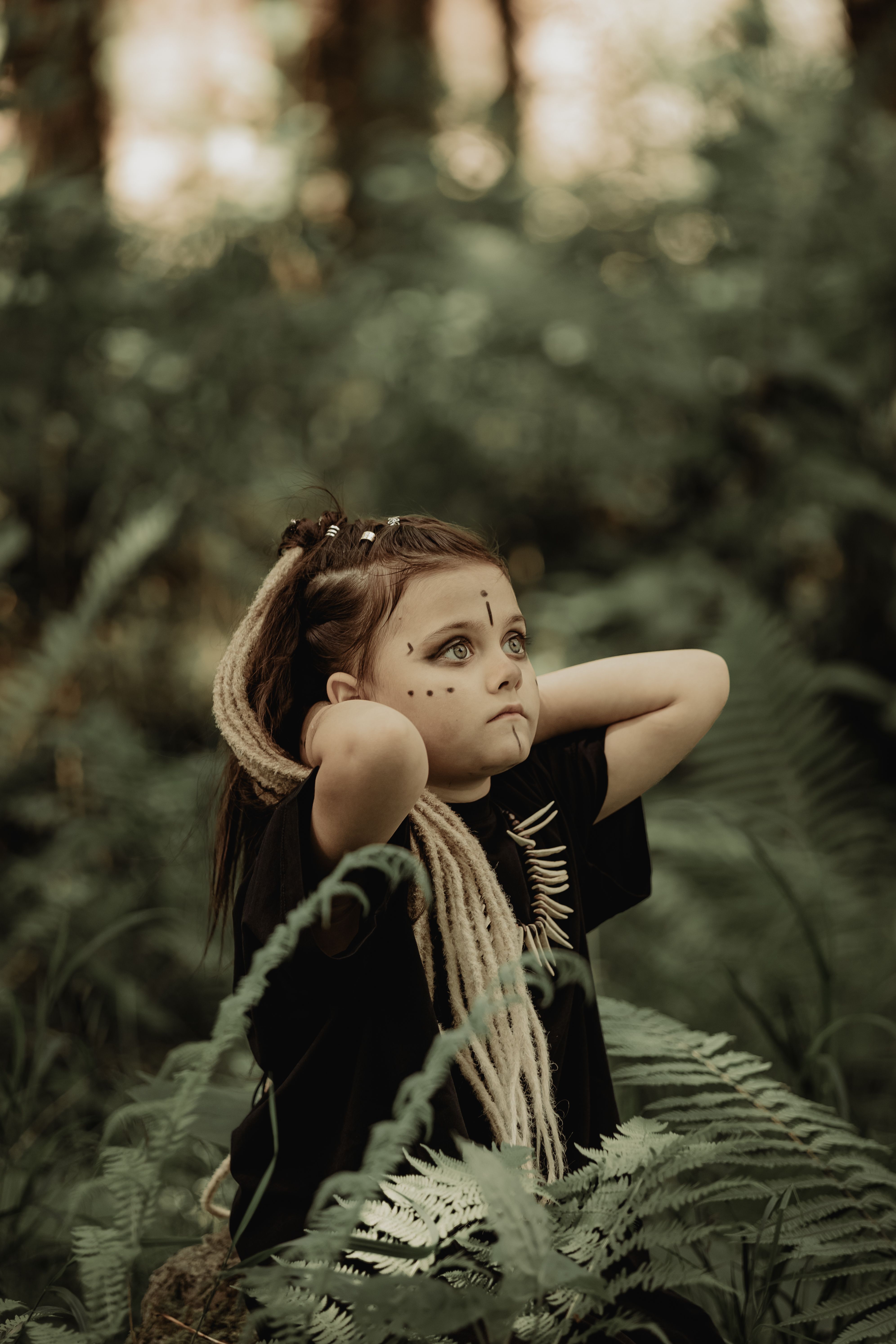 A child with tribal makeup dressed in costume stands in a lush forest setting, looking upwards.