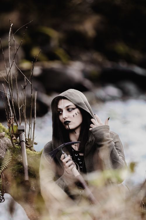 Free Gothic teenager in hooded attire with face art holding axe in a wooded, mysterious setting. Stock Photo