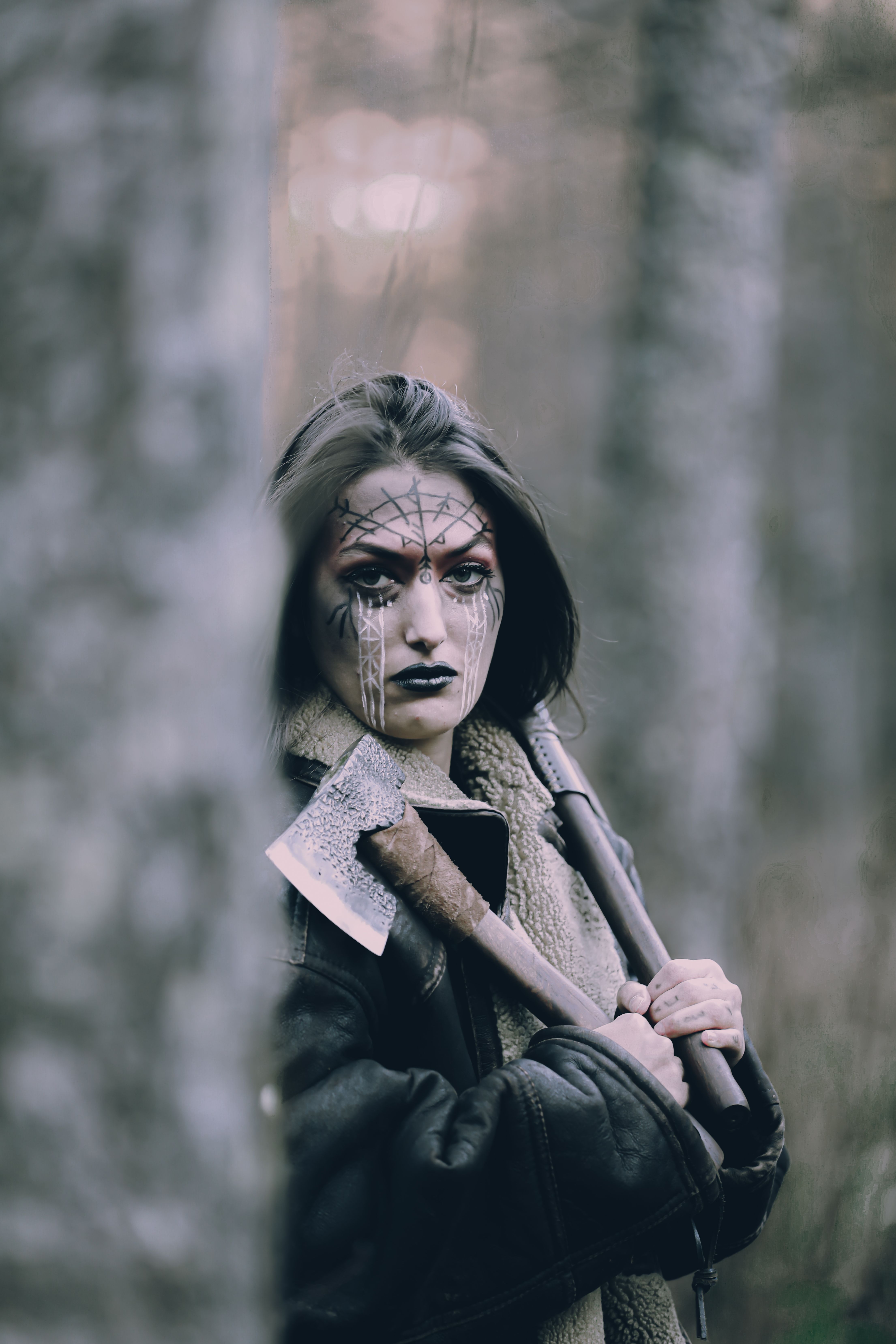 A woman with intricate face paint holds an axe in a misty forest, evoking mystery.