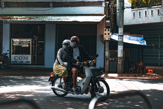 A pair wearing masks and helmets ride a scooter in a sunlit urban setting.