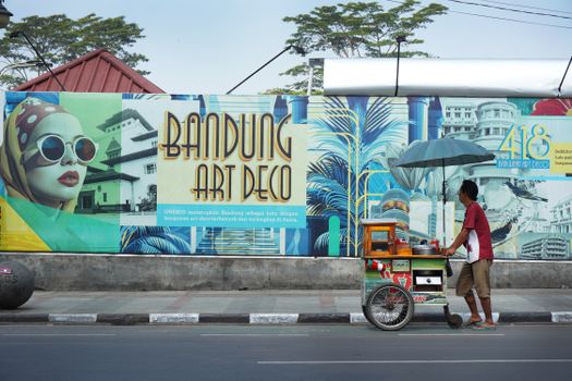 Street vendor with cart passing Bandung Art Deco mural in West Java, Indonesia. - Fotografer Sidoarjo