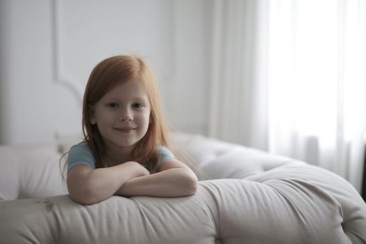 Portrait of a young girl with red hair smiling while leaning on a sofa in a bright room.