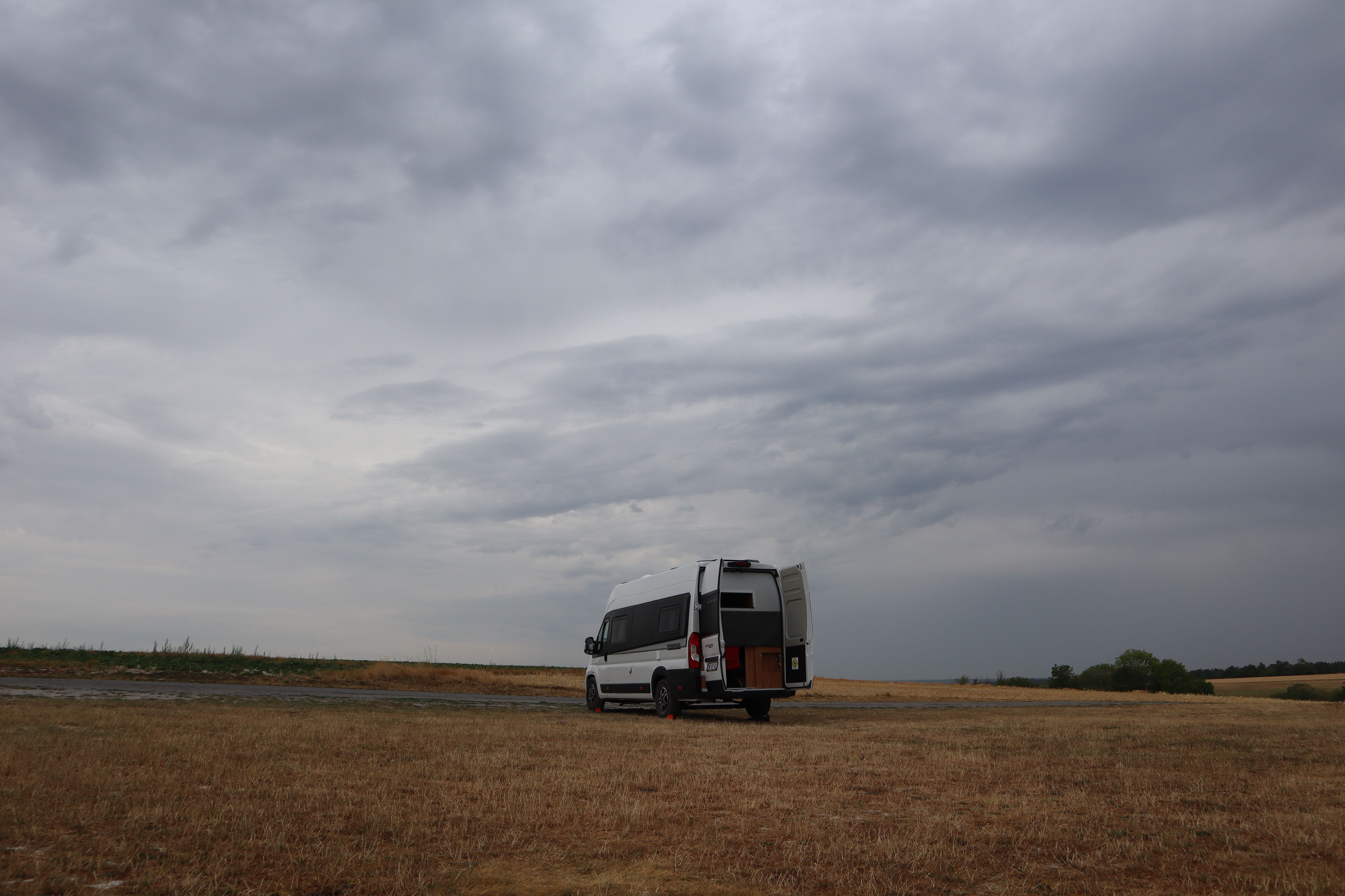 Camper van parked in open farmland under a dramatic, cloudy sky in Massiges, France.