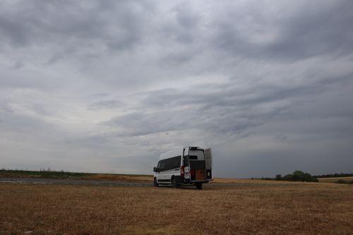 Free Camper van parked in open farmland under a dramatic, cloudy sky in Massiges, France. Stock Photo