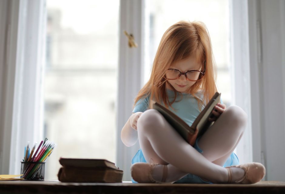 A young girl with glasses reads a book by a window, embodying focus and learning.