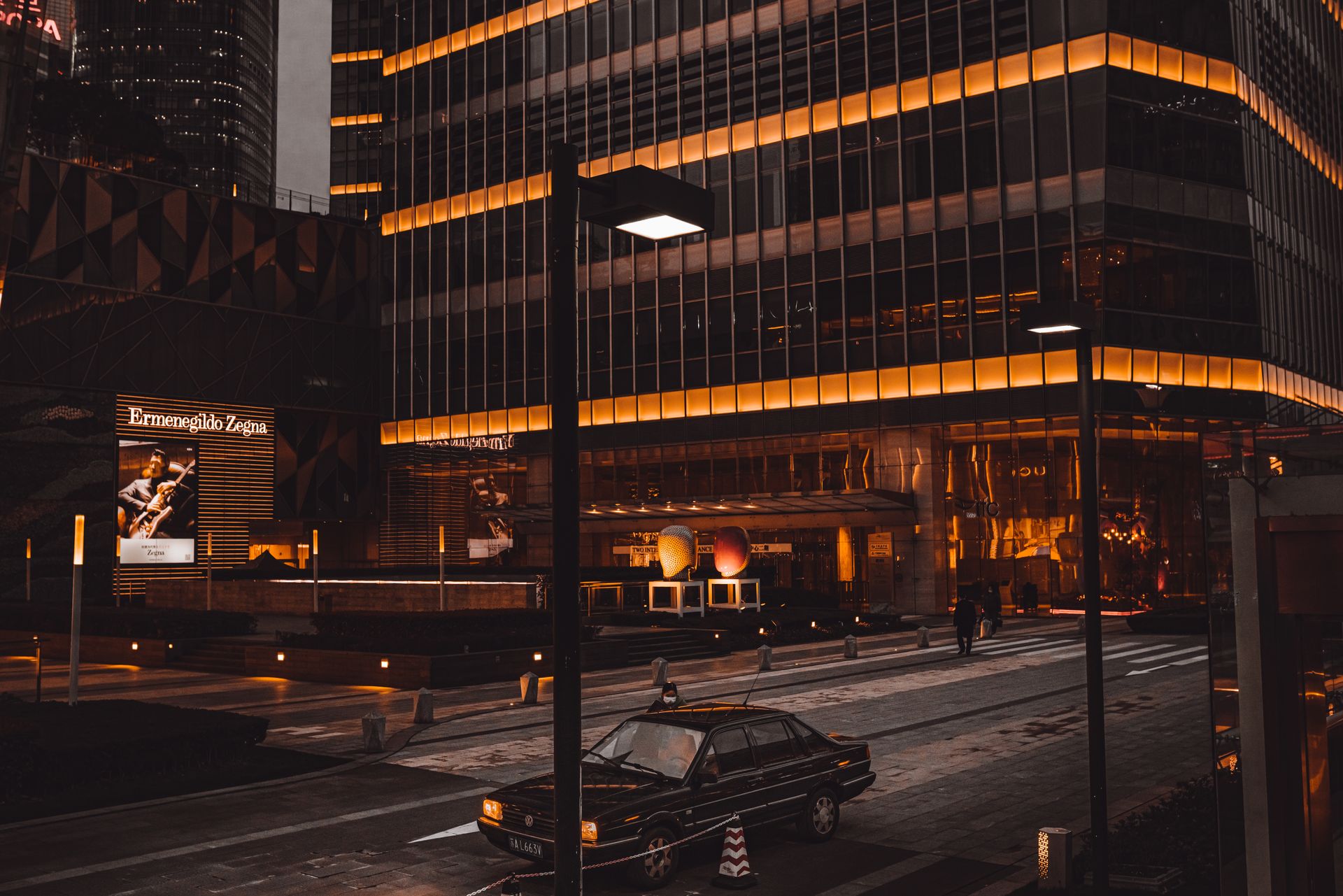 A bustling city street at night featuring an illuminated skyscraper facade and parked vehicle.