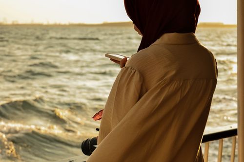 Free A woman in a hijab views the ocean, holding a smartphone during sunset. Calm and serene vibe. Stock Photo