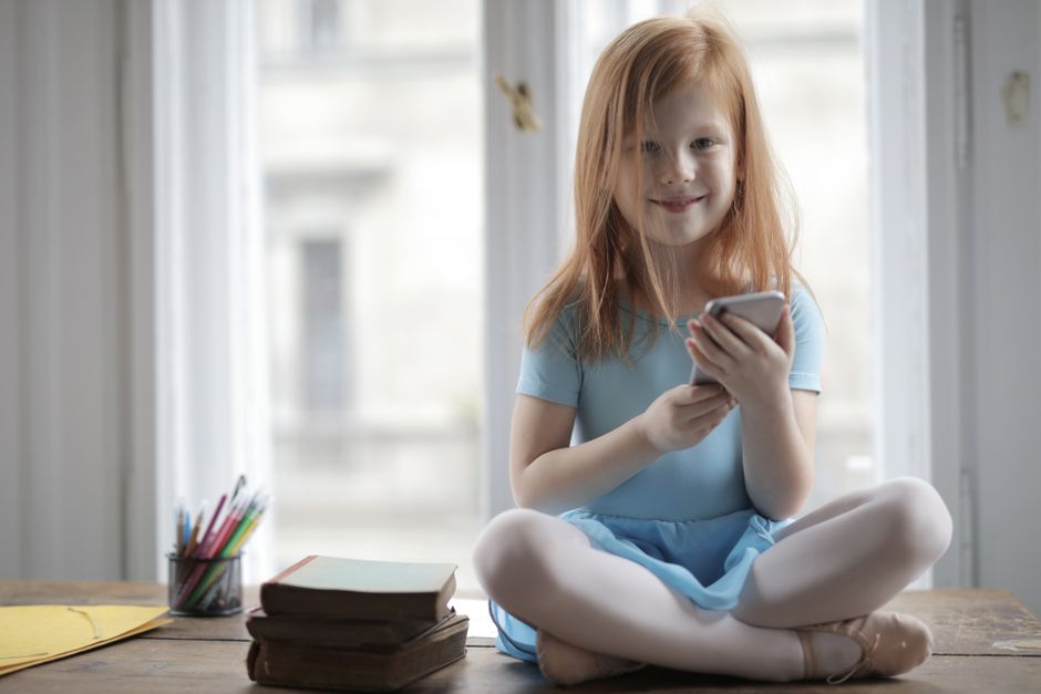 Photo by Andrea Piacquadio on Pexels A cheerful young girl in a blue dress sitting indoors by a window holding a smartphone. - Photo by Andrea Piacquadio on Pexels