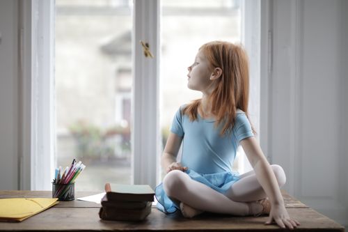 Free Cute red haired peaceful kid wearing blue ballet dress and pointe shoes sitting with crossed legs on wooden desk with books and office supplies against window and looking away dreamily Stock Photo