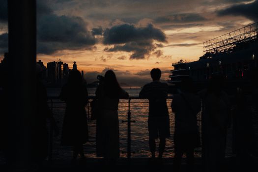 Silhouetted figures admire a dramatic sunset over Hong Kong harbor, capturing urban and natural beauty.