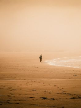 A lone person strolls along a misty beach at sunset, evoking solitude.