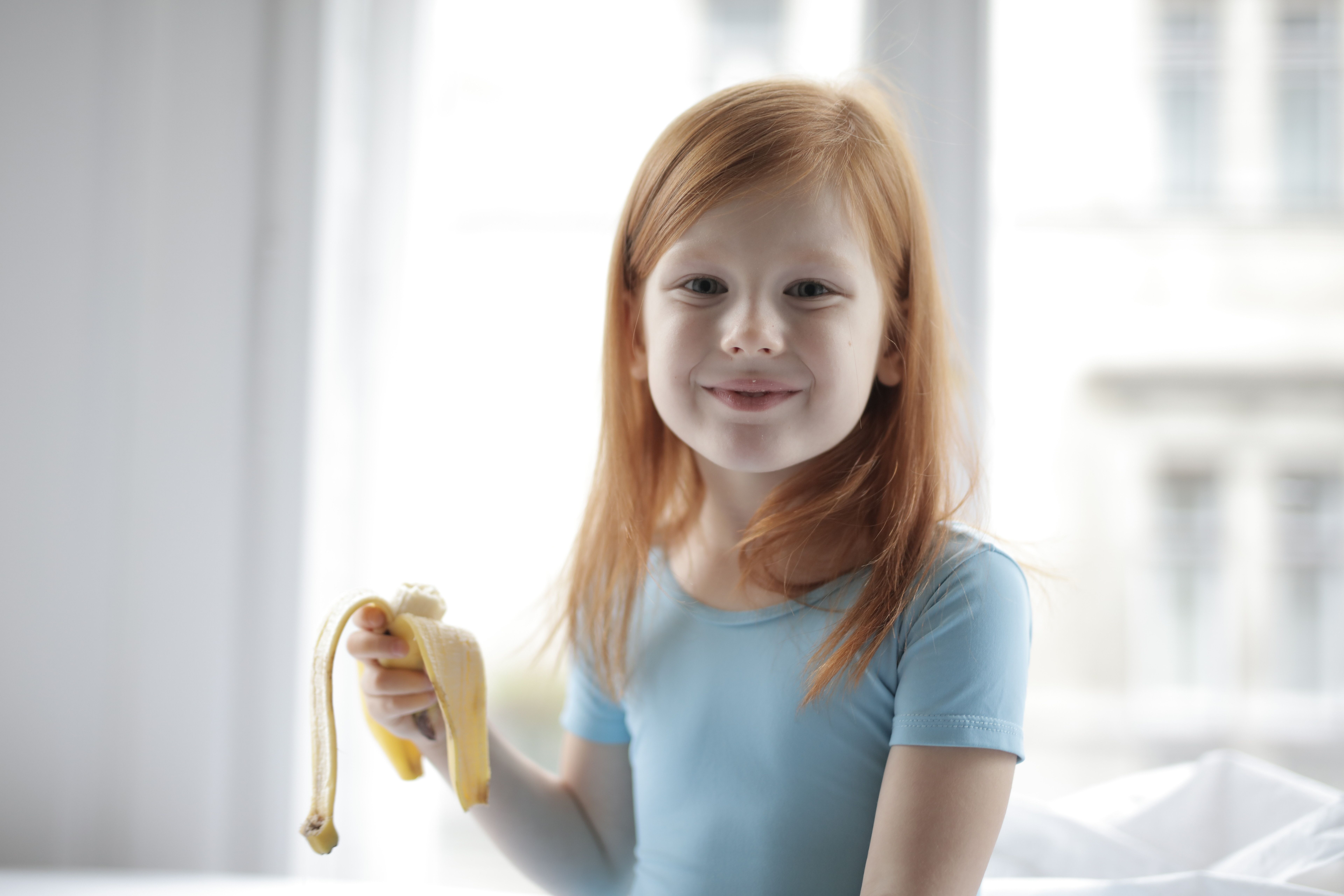 Young girl in a blue shirt holding a peeled banana, smiling indoors.