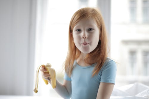 Adorable red haired kid wearing blue casual clothes enjoying taste of fresh banana in light bedroom in morning while looking at camera happily