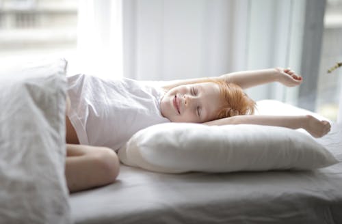 Little adorable child with red hair lying in bed with white linen while trying waking up early morning stretching hands after sleep in light cozy bedroom at home