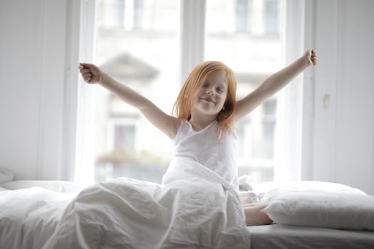 A happy child with red hair stretches on a bed, bathed in soft morning light by a window.