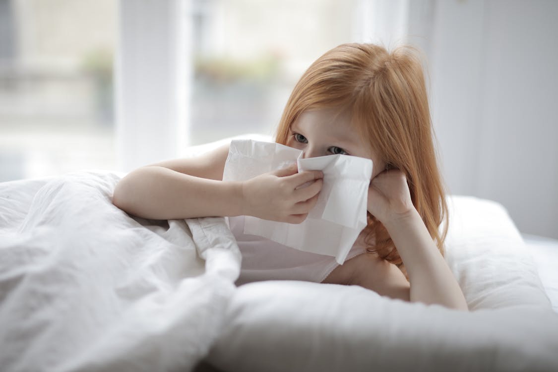 Cute unhealthy child with red hair blowing nose with paper towel while lying on bed with white linen in cozy light bedroom