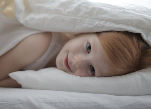 Adorable glad child lying in warm bed with white linen under duvet and looking at camera with happy smile waking up in morning