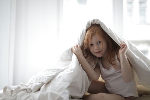 Free A cute young girl with red hair playfully hiding under a blanket in a bright bedroom. Stock Photo