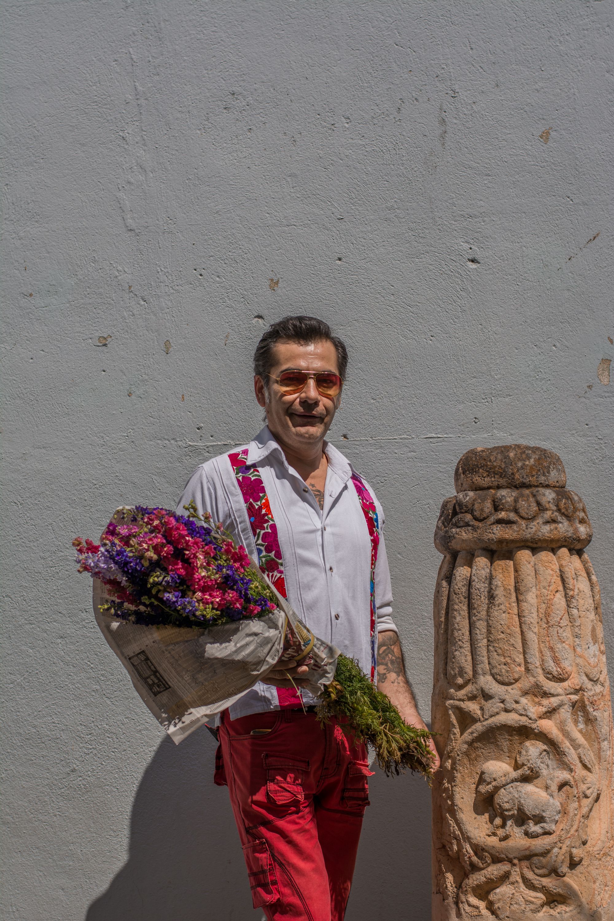 Man in Oaxaca holding colorful bouquet against a rustic wall, wearing casual attire and sunglasses.