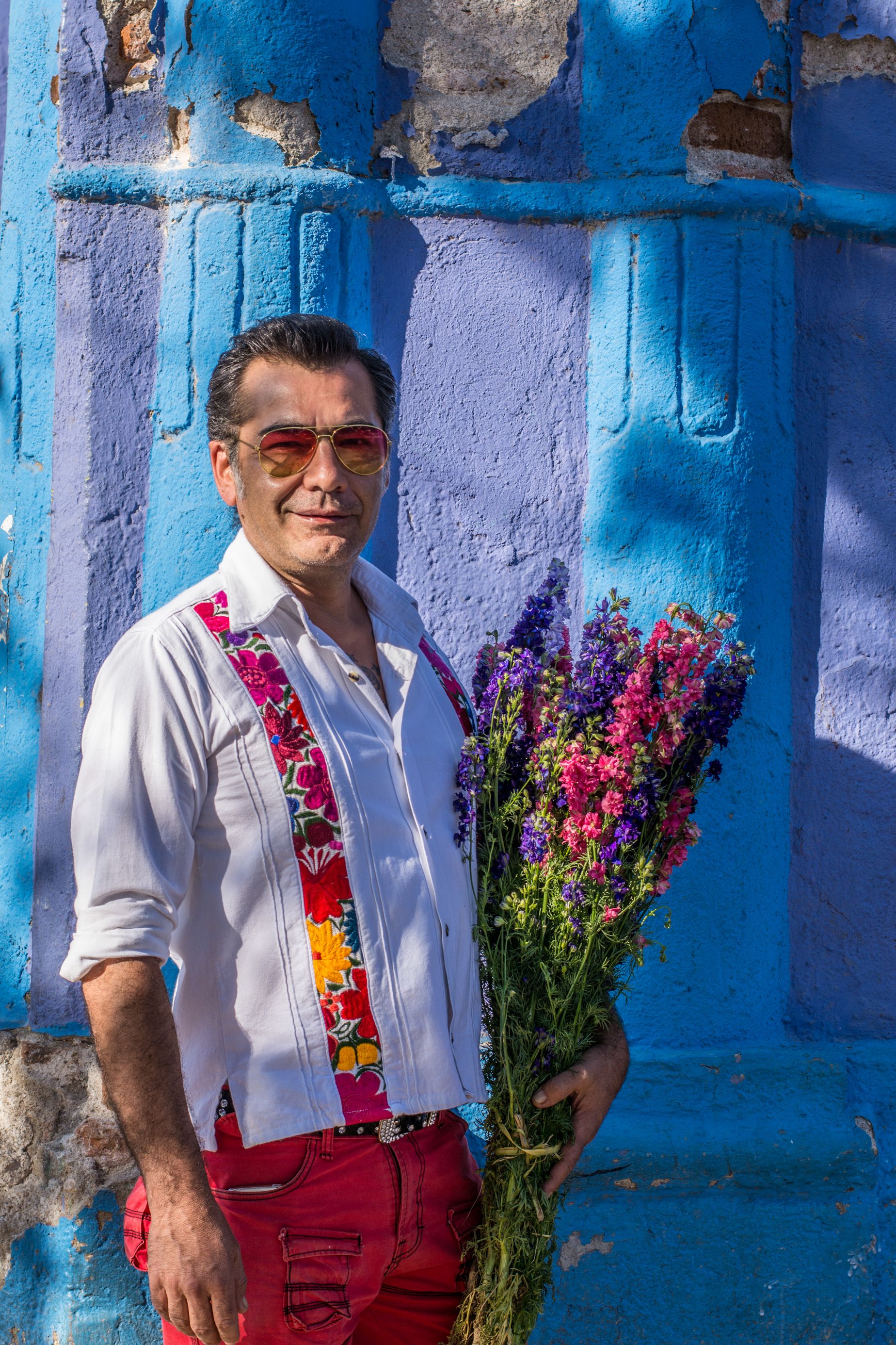 Man in Oaxaca holding flowers against blue wall, showcasing local culture and vibrant colors.