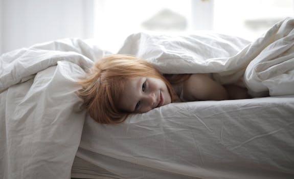 A smiling child with red hair lying comfortably under a white blanket, enjoying a cozy morning in bed.