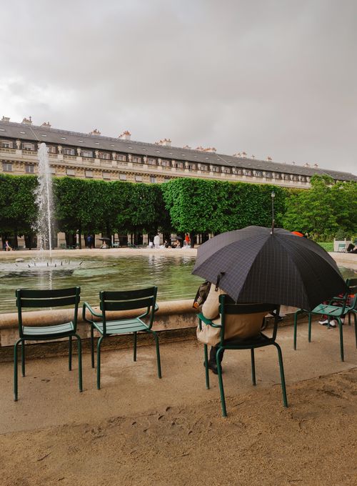 Free A person sits under an umbrella by a fountain in a Paris park, creating a tranquil rainy scene. Stock Photo