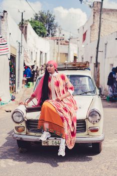Fashionable woman in colorful attire sitting on a classic car in a vibrant city street.