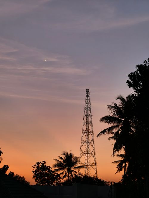 Free A serene sunset sky with crescent moon, silhouetted palm trees, and a transmission tower. Stock Photo