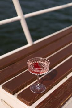 A crystal wine glass with red liquid placed on a boat deck with water in the background.