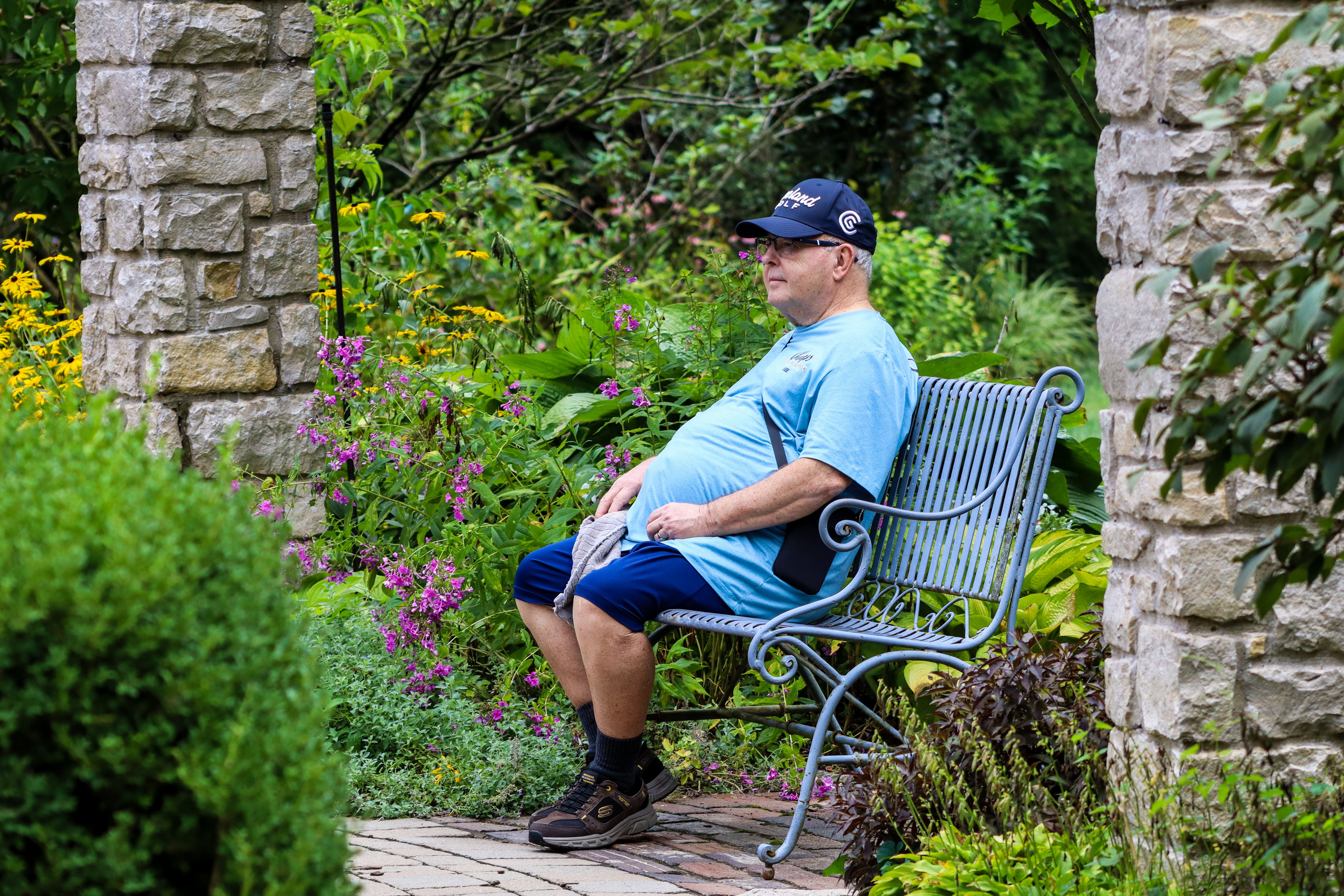 Older man seated on a garden bench surrounded by lush greenery and flowers, enjoying a peaceful summer day.