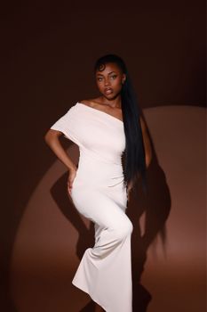 Stylish woman in a white dress poses gracefully against a warm brown backdrop.