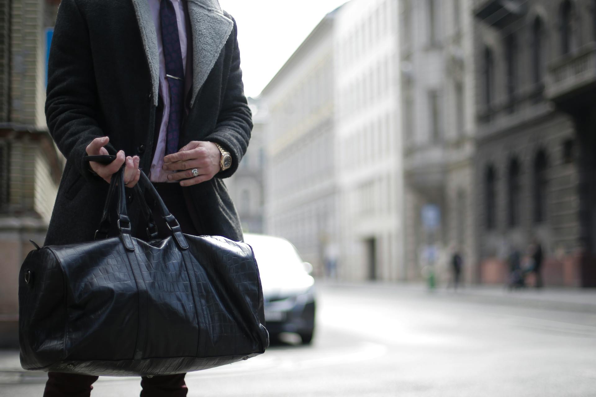 Man in a trench coat holding a travel bag on an urban street, showcasing fashion and lifestyle.