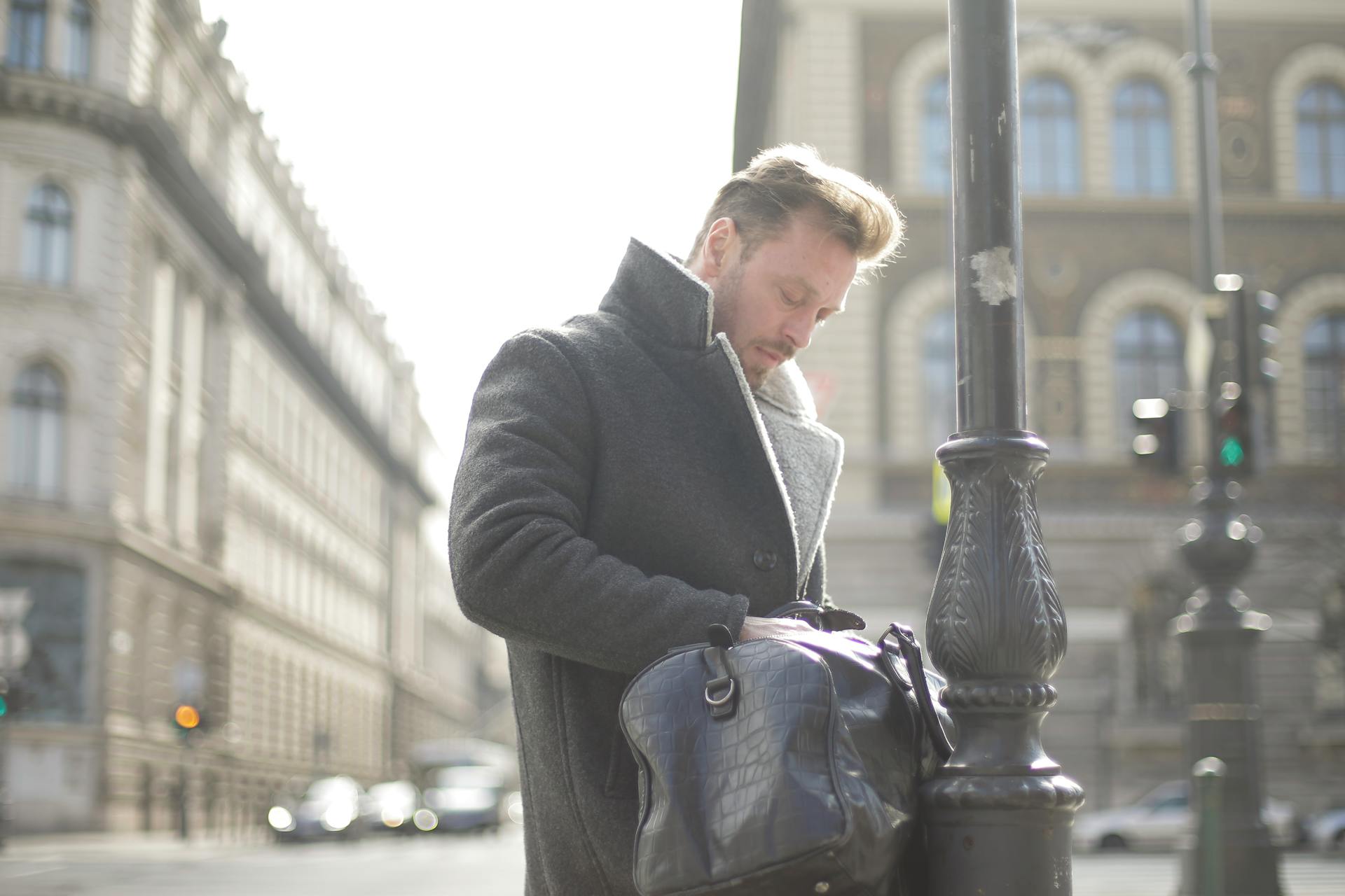 Concentrated handsome bearded male traveler wearing sheepskin coat looking something inside bag for travel standing near retro lamp post on urban square with classic buildings during daytime in cold season