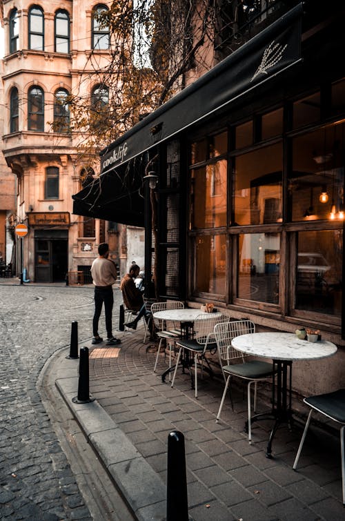 Facade of cafe with large windows and tables on sidewalk along road and faceless people resting in city