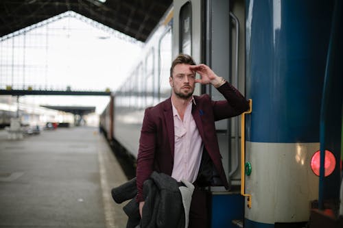 Thoughtful male passenger wearing formal outfit carrying warm coat with hand near head while standing near train with opened door on platform of railroad station during business trip