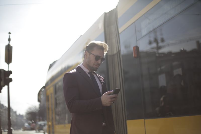Business professional checks phone at a sunny train station, wearing a suit and sunglasses.