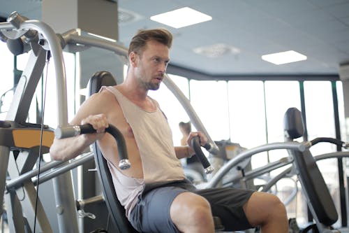 Focused adult male athlete in sportswear using weight machine for arms and chest during workout in modern spacious fitness center in morning