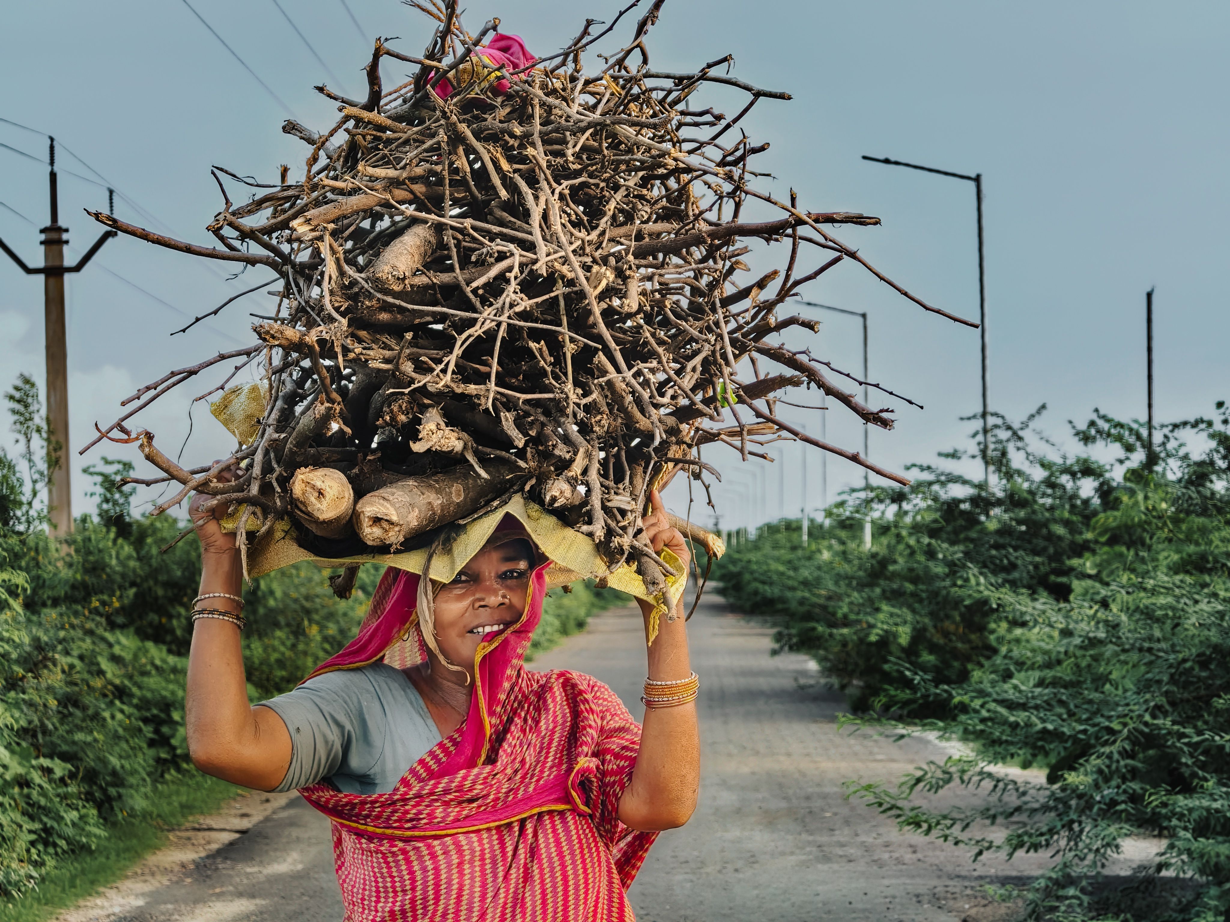 Woman in traditional attire walking on a rural road with firewood on her head.