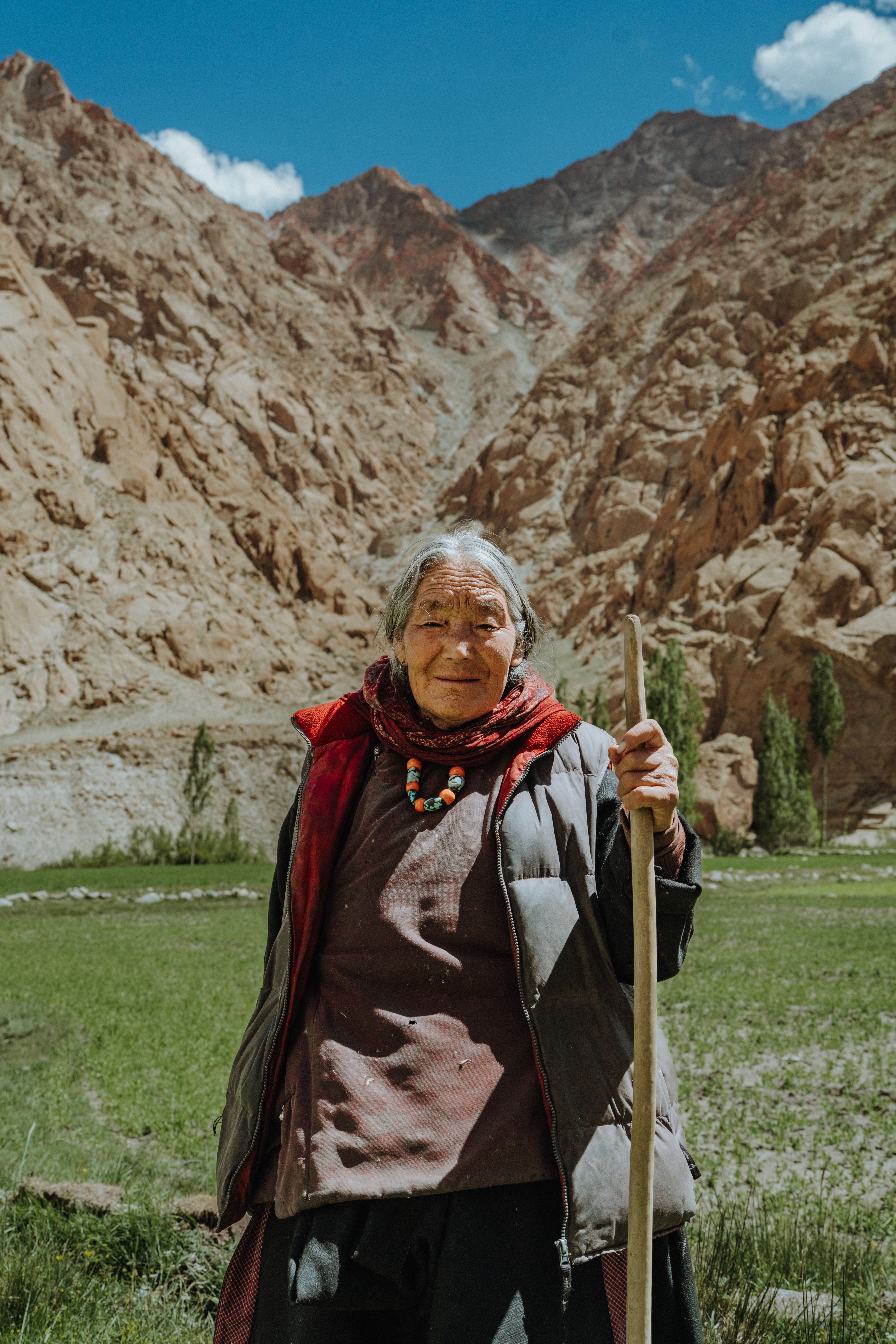 Elderly woman stands confidently in a beautiful mountainous terrain under a clear blue sky.