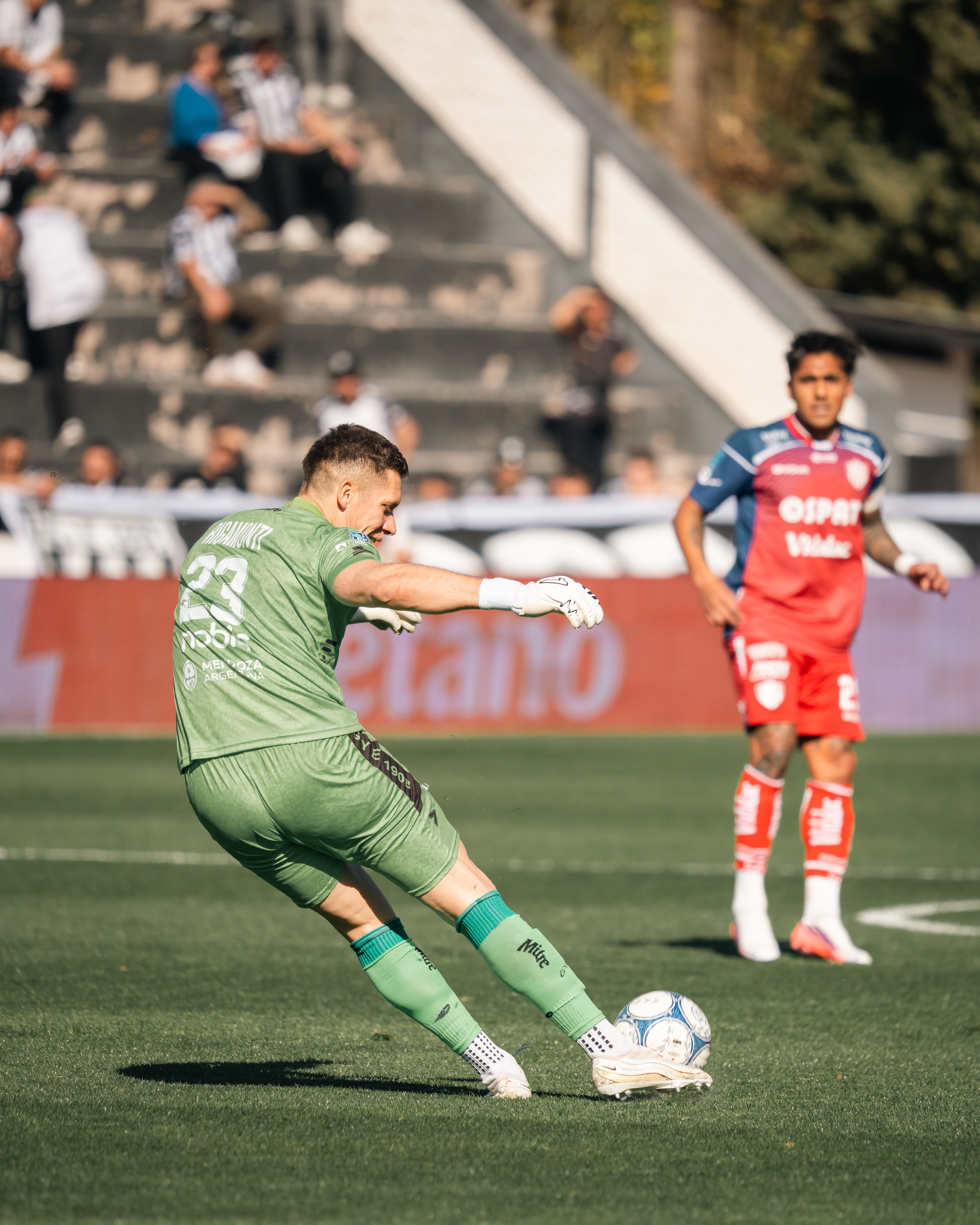 Goalkeeper in green gear kicks the ball during a sunny soccer match with players and audience in background.