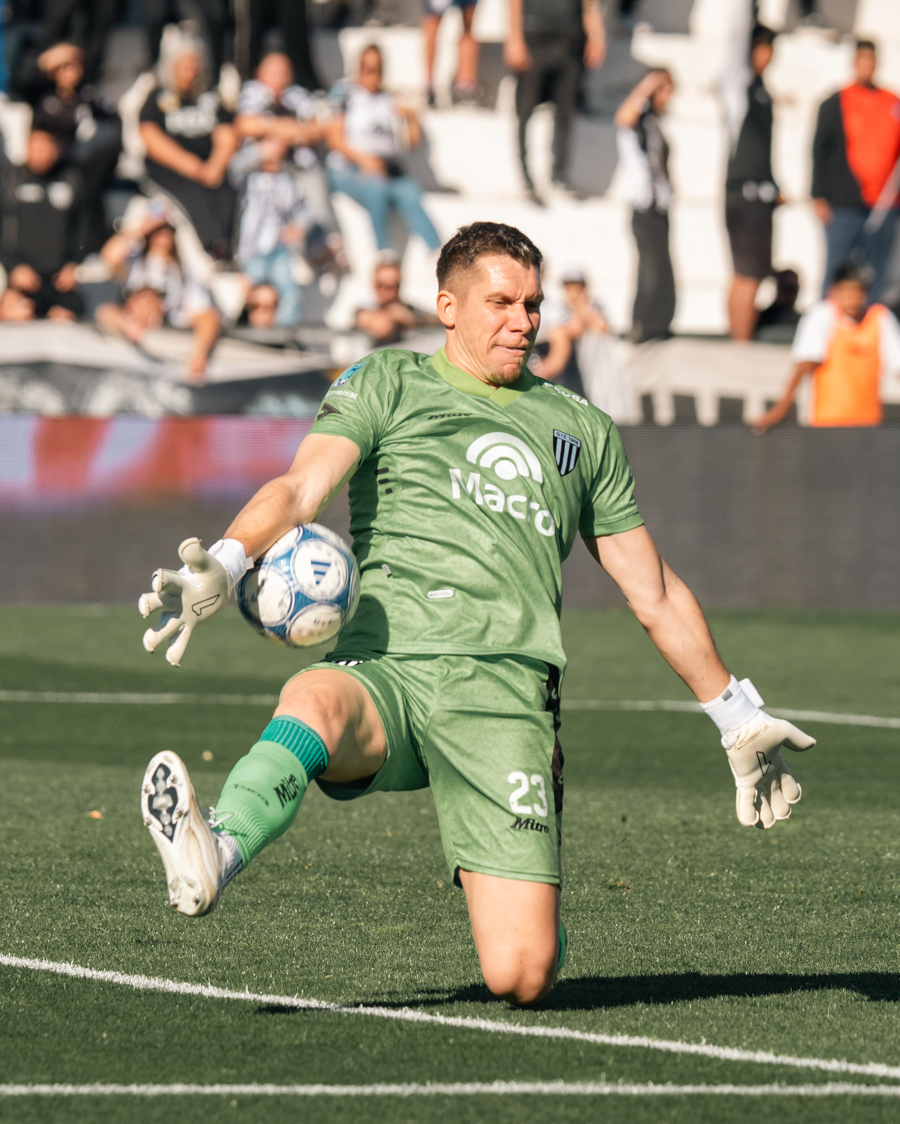 Professional goalkeeper in green uniform kicking a soccer ball during an outdoor match.