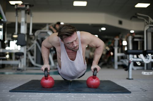 Adult male athlete wearing activewear performing push up exercise with kettlebells during fitness workout on floor of modern spacious sport club and looking away