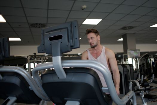Confident muscular adult male athlete in sportswear standing near running machine in modern spacious fitness center before training while looking away pensively