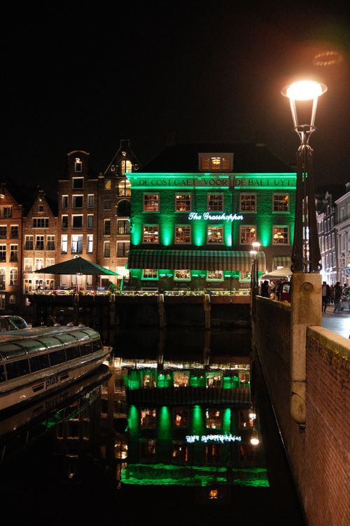 Free A picturesque canal scene in Amsterdam with a green-lit historic building reflected on the water at night. Stock Photo