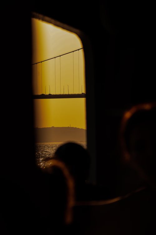 Free Silhouette of a suspension bridge at sunset viewed from a window with a blurred figure in the foreground. Stock Photo