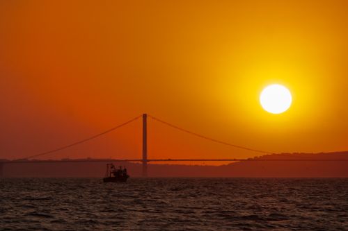 Free Golden sunset silhouette of a suspension bridge with a boat on the sea, creating a breathtaking view. Stock Photo